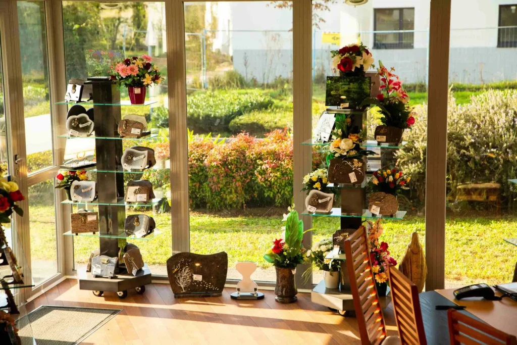 Présentoirs en verre exposant des urnes, plaques funéraires et compositions florales devant une baie vitrée